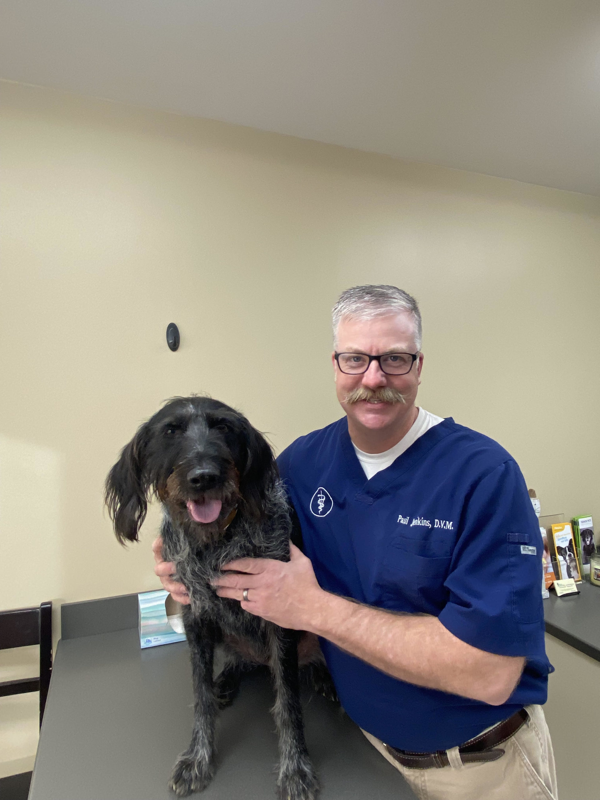 J. Paul Jenkins DVM, veterinarian at Vilonia Animal Clinic in Conway, AR, wearing a blue shirt and khaki pants while holding a large black dog on a table